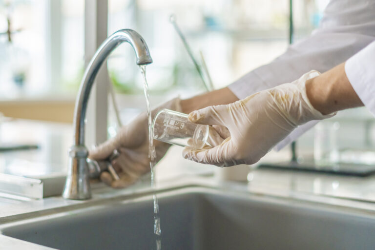 a gloved hand holding a glass beaker under a kitchen faucet, collecting water